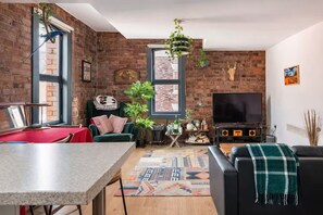 A modern living room with a brick wall, a large window, a flat-screen TV, a sofa, a dining table, and various plants.