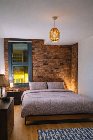 A bedroom with a brick wall, a bed with patterned bedding, a hanging light fixture, and a window with blinds.