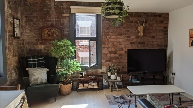 A living room with a brick wall, a large window, a television, a sofa, a coffee table, and a potted plant. - Cozy 2-bedroom apartment in welcoming Liverpool with WiFi (Liverpool)