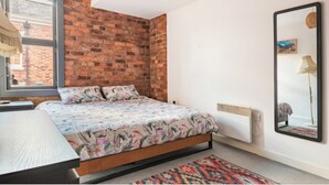 A bedroom with a brick wall, a bed with floral bedding, a bedside table, a radiator, a mirror, and a window with a view of a building. - Cozy 2-bedroom apartment in welcoming Liverpool with WiFi (Liverpool)