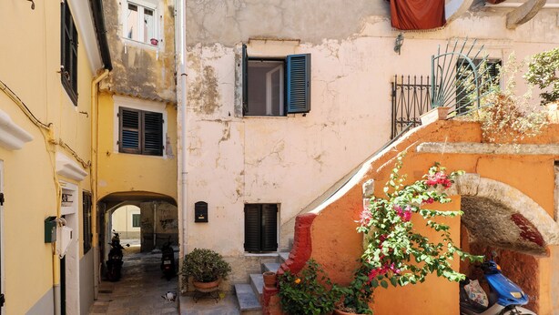 Une rue étroite où est garé un scooter devant un immeuble, avec un pot de fleurs et une petite arche.