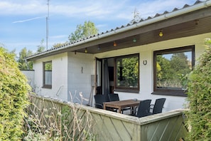A single-story building with a patio area featuring a table and chairs, a window with a view of greenery, and a door.