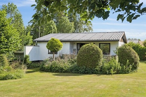 A small, single-story house with a tiled roof, surrounded by a well-maintained lawn and dense foliage.