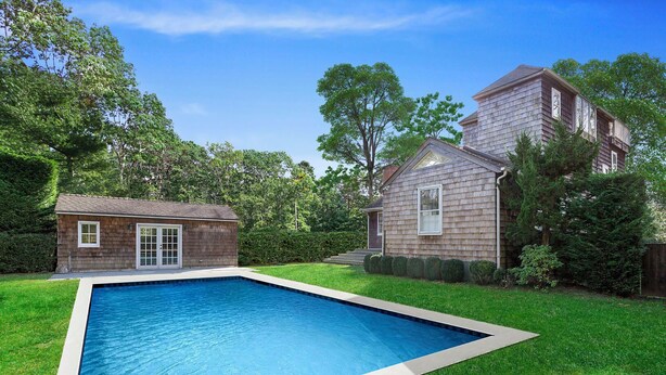 A rectangular swimming pool with a white border, surrounded by a well-maintained lawn and a wooden house with a garage. - 4-bedroom house in charming Wainscott walking distance to the beach (East Hampton)