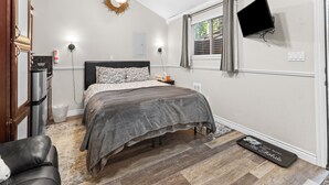 Une chambre à coucher avec un lit, un téléviseur fixé au mur, une fenêtre avec des rideaux, une porte et un petit tapis.
