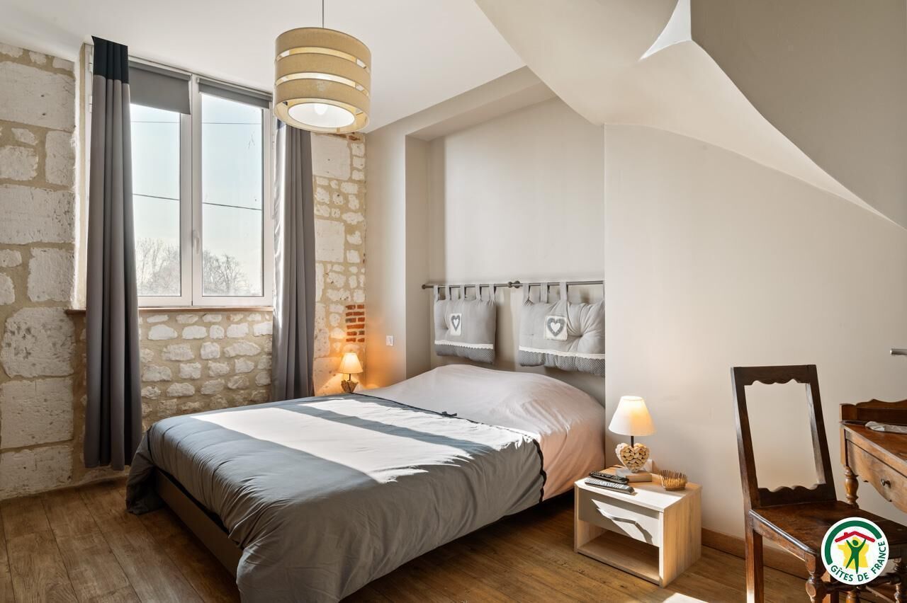 Ein Schlafzimmer mit Bett, Nachttisch, Lampe, Spiegel und einem Fenster mit Vorhängen.