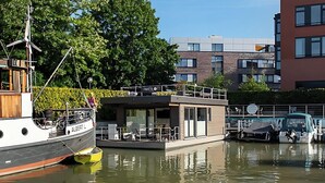 Una moderna casa flotante amarrada en un puerto deportivo, con zona de comedor y una pequeña terraza.