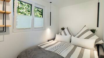 A bedroom with a bed, striped bedding, a bedside table with a plant, and a window with blinds.