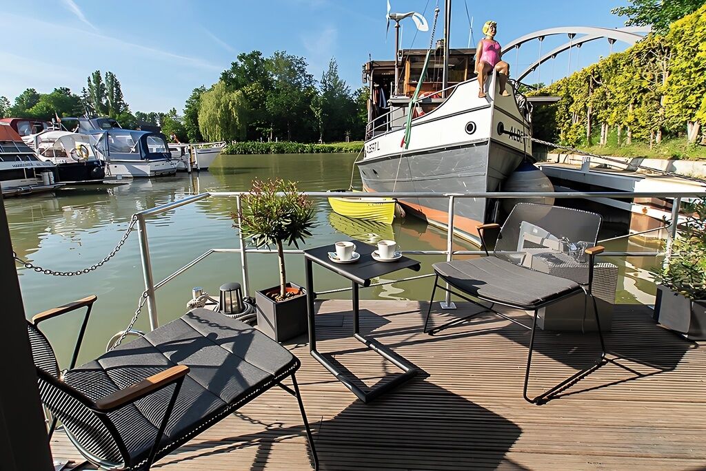 Um terraço com área de estar externa, uma pequena mesa com xícaras e vista para um cais com barcos.