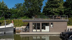 Una moderna casa flotante atracada en un puerto deportivo, con una cubierta techada y una sala cerrada con vidrio.