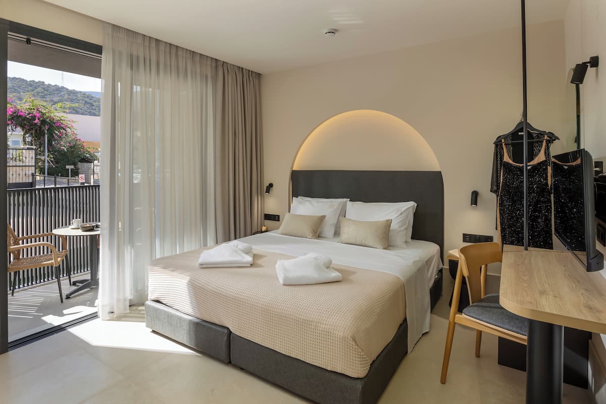Habitación de hotel con una cama grande, una mesa de madera, una silla y un balcón con vistas a las colinas.