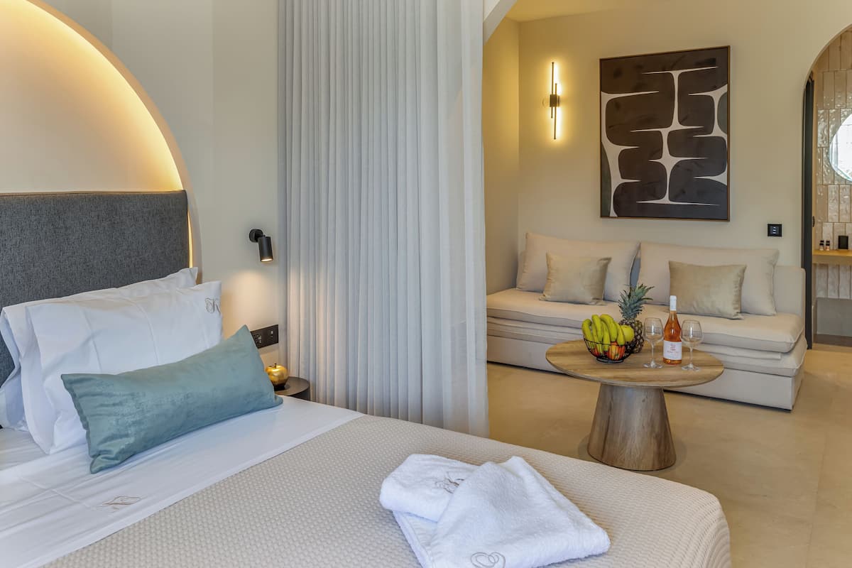 Habitación de hotel con cama, sofá, una mesa de centro con fruta y una botella, y una lámpara de pared.