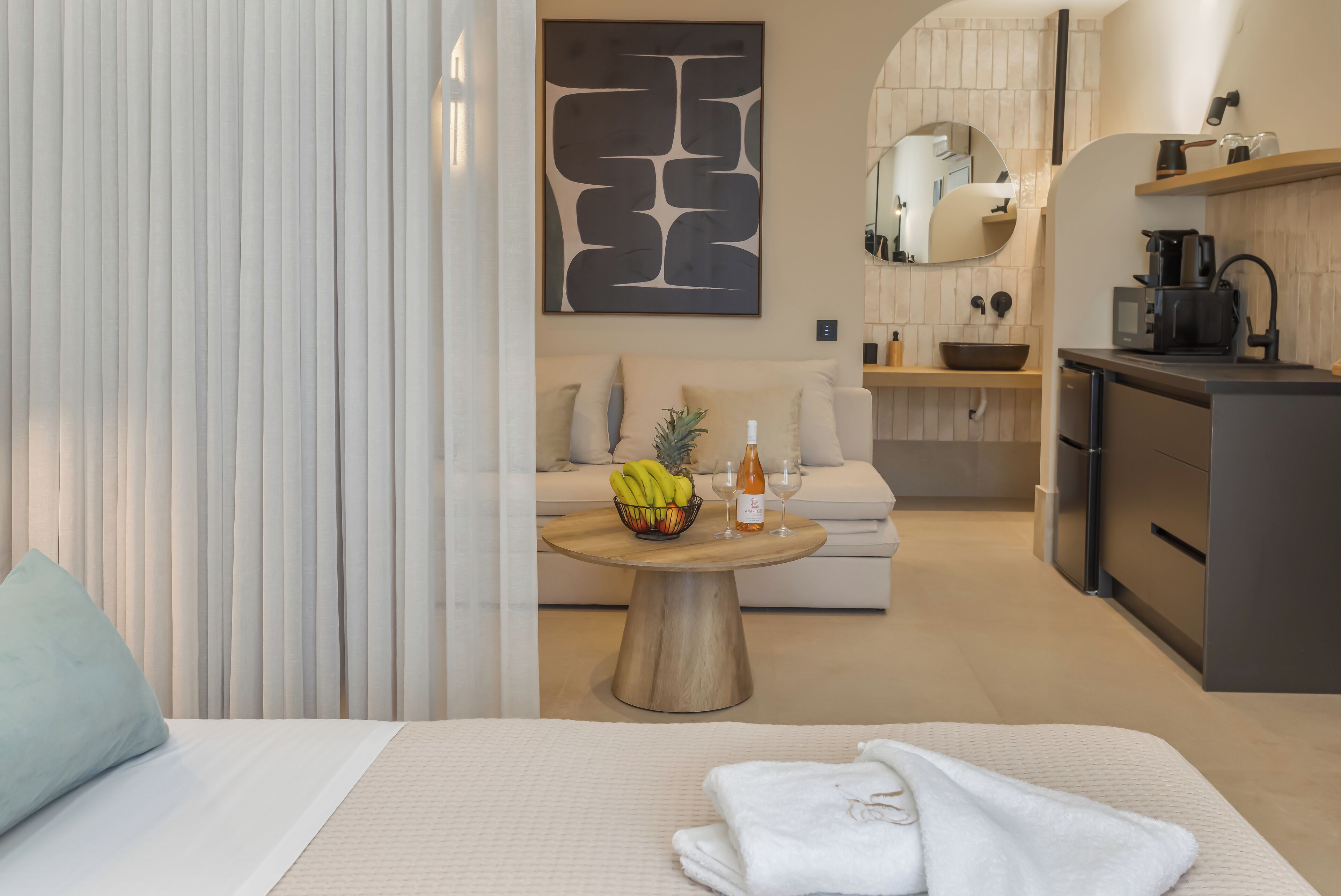 A hotel room with a bed, a dining table with fruit and a bottle, a kitchen area, and a living space with a sofa and a decorative wall piece.