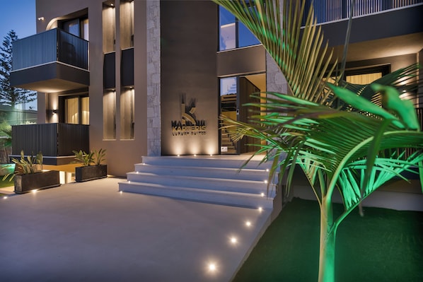 A modern building with a sign reading "KARERIIS LUXURY SITES," featuring a staircase leading to the entrance, a balcony, and a well-lit exterior. - Karteris Luxury Suites (Stalida)