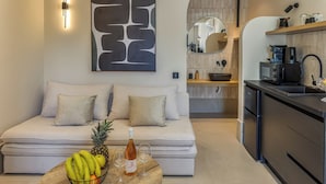 Een moderne woonkamer met een bank, een bijzettafeltje met fruit en een fles, en een keukenhoek met apparatuur en een spiegel.