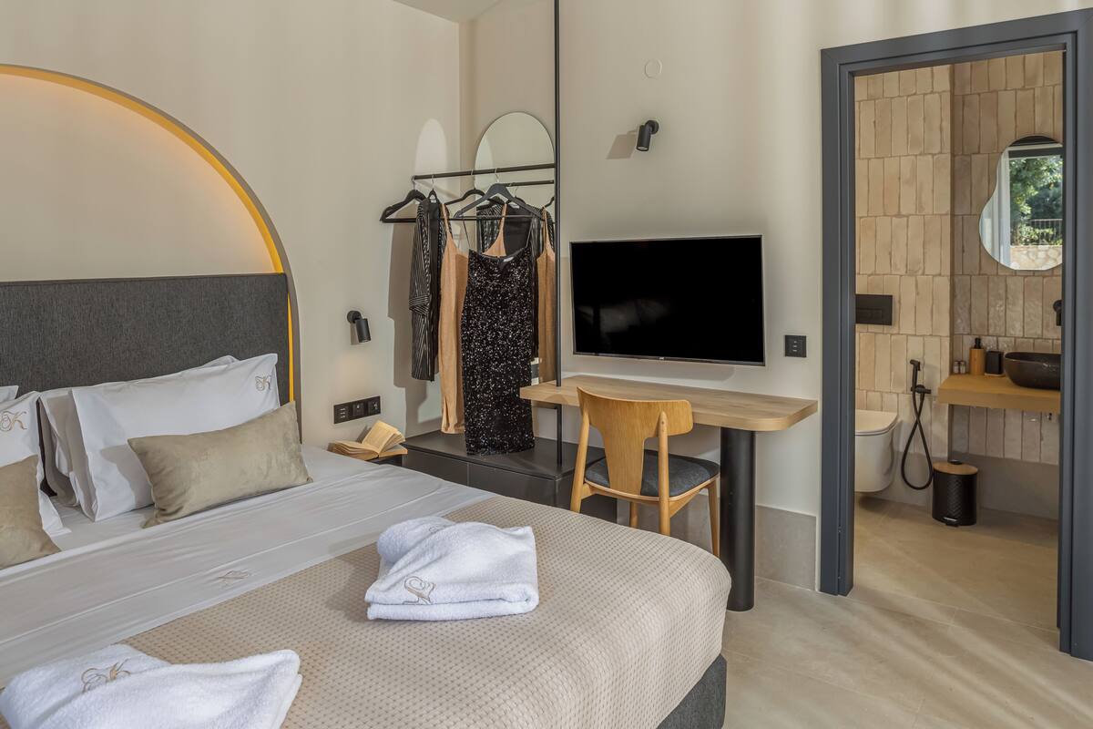 Habitación de hotel moderna con cama, escritorio, televisión, espejo y baño visible a través de una puerta abierta.