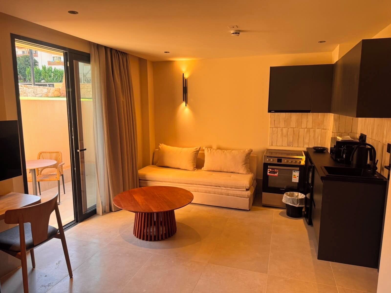 A compact hotel room with a sofa, a small dining table, a kitchenette, and a sliding glass door leading to an outdoor area.
