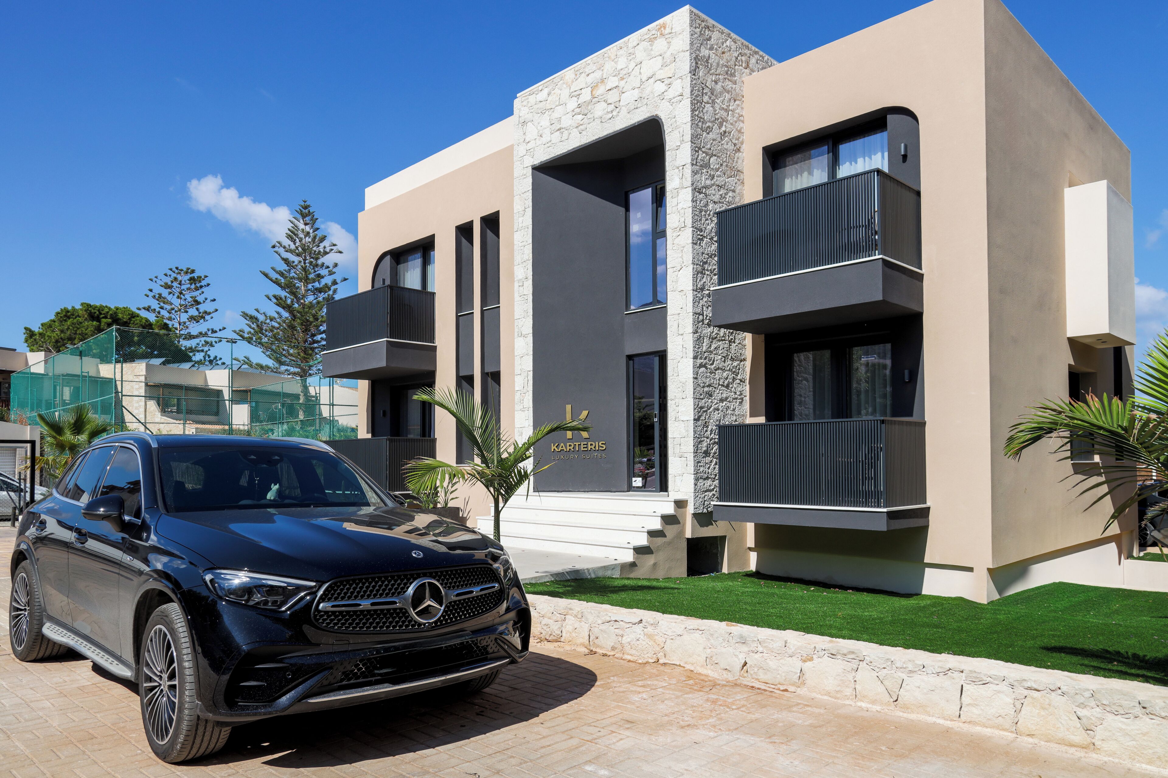 A modern building with a stone facade, a Mercedes-Benz SUV parked in front, and a sign indicating 'KARTEKIS LUXURY SUITES'.