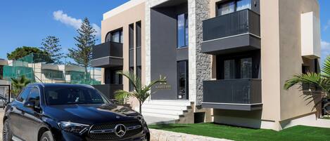 A modern building with a stone facade, a Mercedes-Benz SUV parked in front, and a sign indicating 'KARTEKIS LUXURY SUITES'.