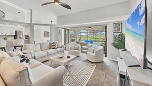 Un ampio soggiorno con un grande divano, tavolino e televisore. Si nota una ventola a soffitto, una zona cucina con mobili bianchi e vista su una piscina e un'area esterna.