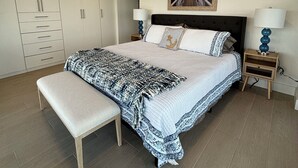 Un dormitorio con una cama grande, un banco, dos mesitas de noche, un armario y un cuadro en la pared.
