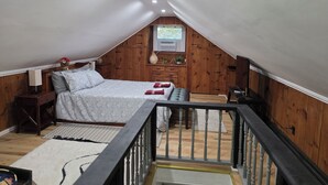 Une chambre avec un plafond en pente, des murs en bois, un lit avec une literie à motifs, une table de chevet, une commode et une fenêtre donnant sur un paysage verdoyant.