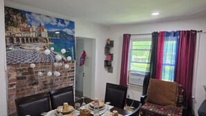 Un coin repas agrémenté d’une fresque représentant une ville côtière, d’un mur en briques et d’une table à manger mise avec des assiettes, des verres et un vase décoratif.