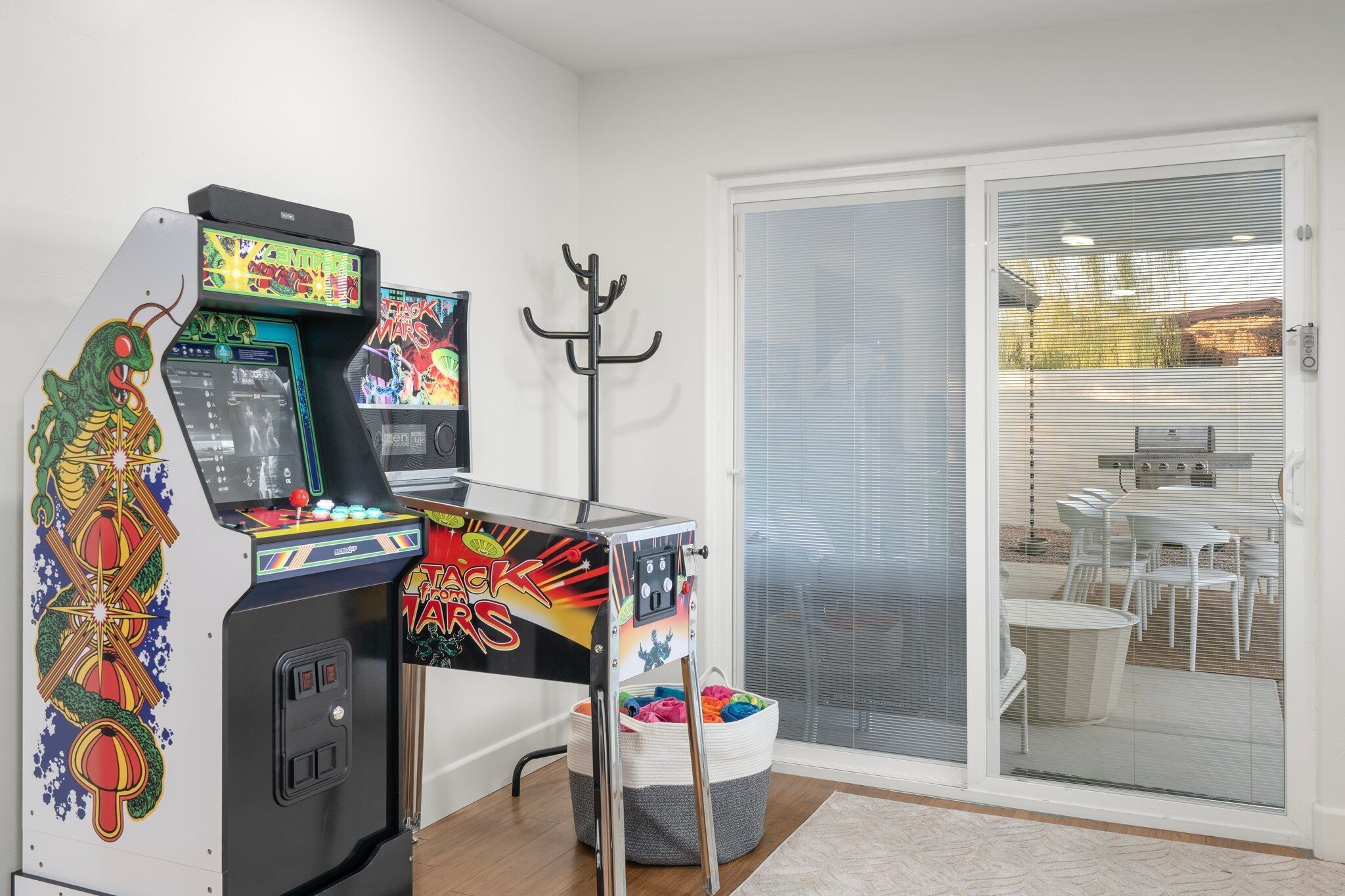 A room with arcade games, a coat rack, and a sliding glass door leading to an outdoor patio with chairs.
