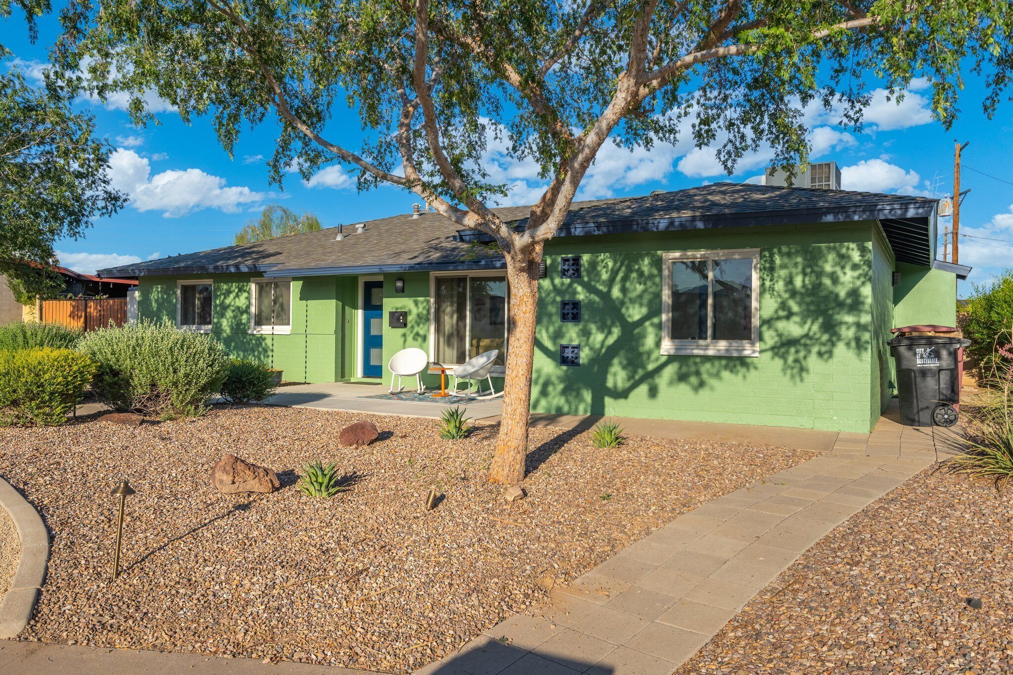 A green house with a covered patio, a tree, and a gravel driveway.