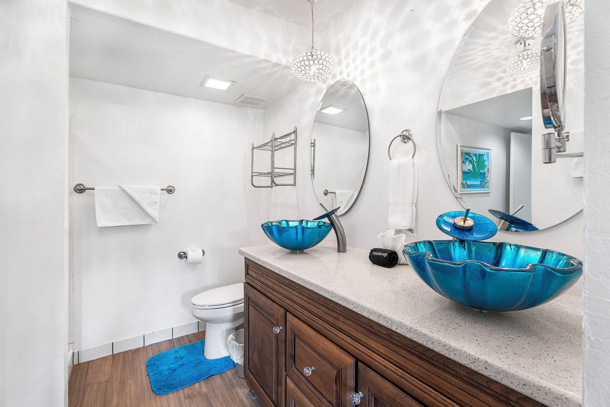 Une salle de bain moderne avec des toilettes blanches, un meuble-lavabo en bois doté d’un vasque bleue et un grand miroir au-dessus du lavabo.