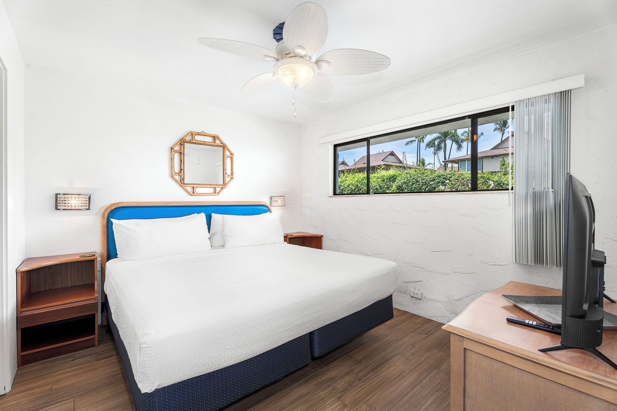 A hotel room with a bed, a TV, a ceiling fan, a window with a view of palm trees, and a wooden nightstand.