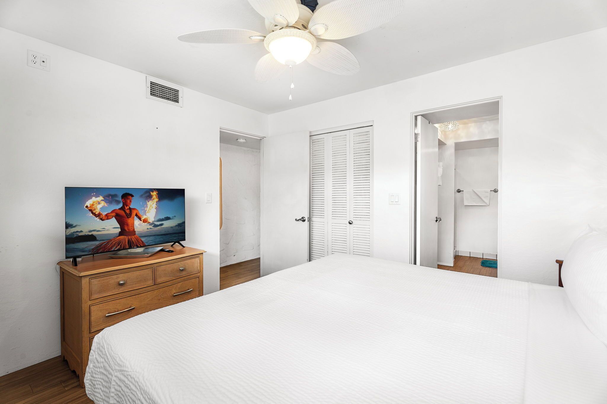 Une chambre avec un grand lit, un ventilateur de plafond, une commode en bois sur laquelle est posée une télévision, et des murs blancs.