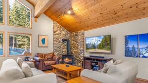 A cozy living room with a stone fireplace, wooden ceiling, and large windows offering views of the outdoors.