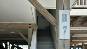 Un escalier en bois mène à une zone abritée, où se trouve un panneau indiquant « B7 ». Une voiture est garée à proximité, et l’entrée est fermée par un portail.