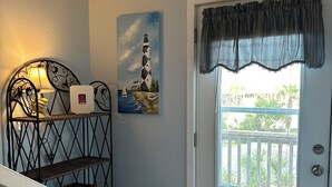Une porte donnant sur un balcon, un tableau représentant un phare, une étagère en métal avec une lampe et un panneau indiquant la direction de Gulf Shores, en Alabama.