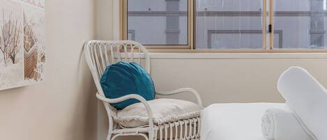 A room with a white wicker chair, a blue cushion, and a white bed with a rolled-up white towel.