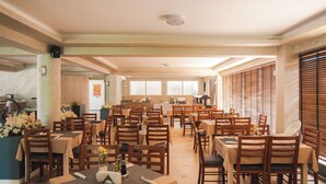 A spacious dining area with multiple tables and chairs, large windows with blinds, and a kitchen area in the background. - EMERALD Hotel (Ulcinj)