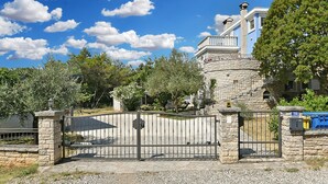 Un portón de piedra con una valla metálica, un edificio de piedra con un balcón y árboles al fondo.