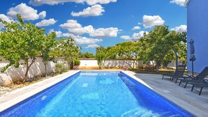Una piscina rectangular con agua azul cristalina, rodeada por una terraza con baldosas. Se observan sillas de descanso y una pérgola con una mesa y sombrilla en el lado derecho.