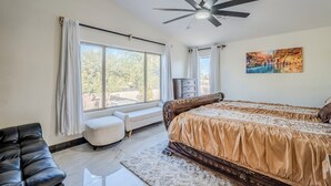 Un dormitorio con una cama grande, ventilador de techo, ventana con cortinas, una banca y un cuadro en la pared.