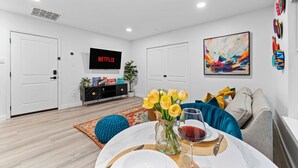 A modern living room with a dining area, a flat-screen TV displaying Netflix, a colorful abstract painting, and a vase of yellow tulips. - Moroccan-Inspired 1-BR Getaway near Reunion Tower (Dallas)