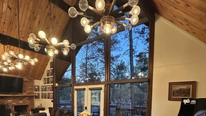 A spacious dining area with a wooden table and chairs, a fireplace, and a large window offering a view of the outdoors. - Out of Office: Cozy A-frame cabin in the pines near trails and adventure! (Munds Park)