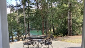 Eine Terrasse mit rundem Tisch und vier Stühlen, umgeben von Bäumen und einem Gewässer.