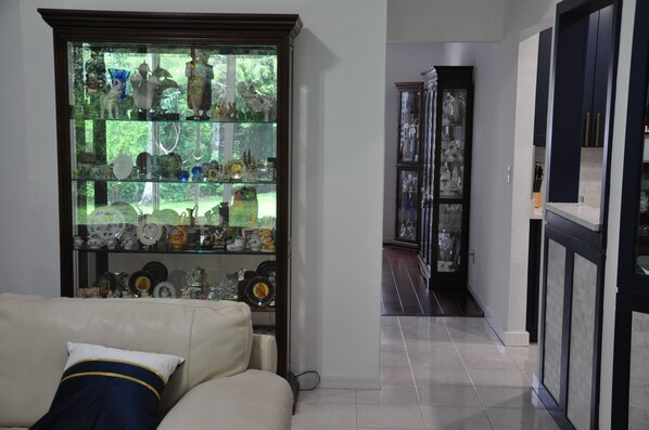 A living room with a beige sofa, a glass display cabinet filled with decorative items, and a hallway leading to a dark-toned room.