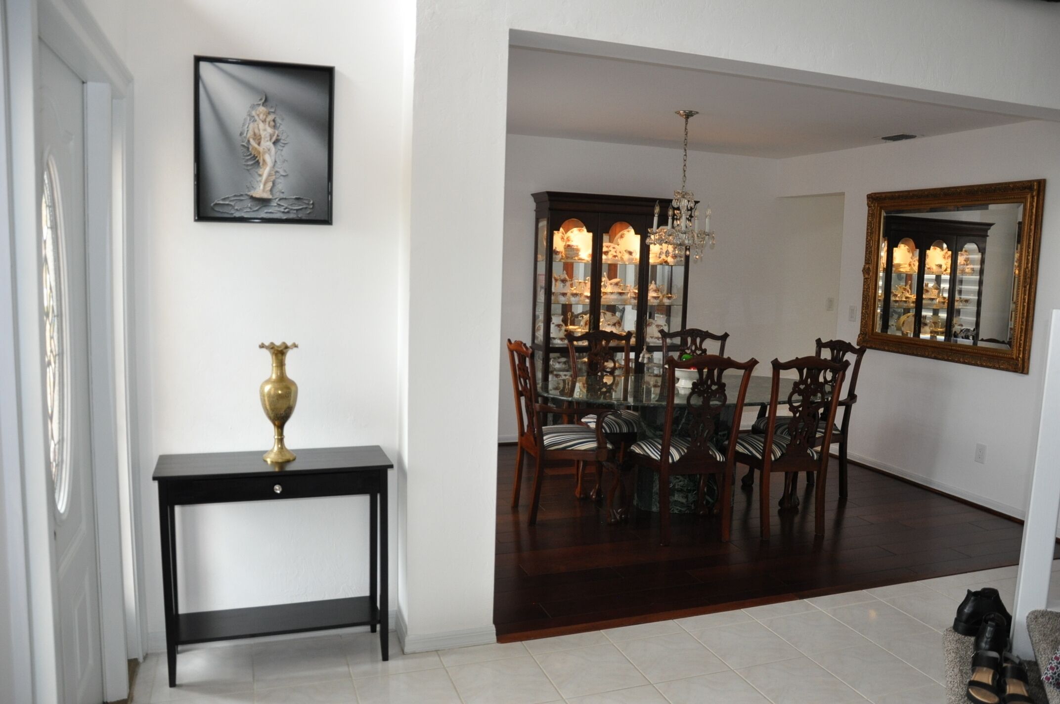 A dining area with a glass-top table, wooden chairs, and a chandelier. There is a black console table with a brass lamp, a framed artwork on the wall, and a large mirror above the table.