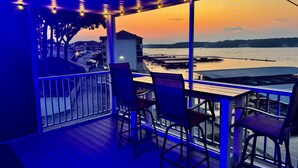 A balcony with chairs and a table, overlooking a waterfront with buildings and a sunset. - Top Rated, Waterfront Condo at 19MM Amazing views! Prime location! Boat slip! (Osage Beach)