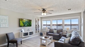 A spacious living room with a large window offering a view of a lake, a ceiling fan, and a grey sofa. - Top Rated, Waterfront Condo at 19MM Amazing views! Prime location! Boat slip! (Osage Beach)