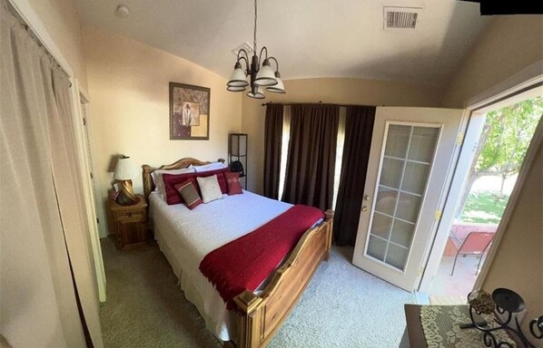 A bedroom with a wooden bed, a nightstand, a lamp, a painting on the wall, and a sliding glass door leading to an outdoor area. - Cozy 1-bedroom apartment, WiFi, AC, Views! Wildlife! In charming Pahrump (Pahrump)