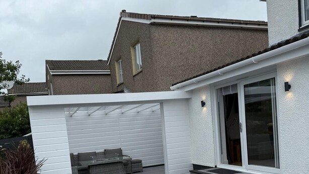 Ein modernes Haus mit überdachter Terrasse, einer Schiebetür und einer kleinen Sitzgelegenheit im Freien.