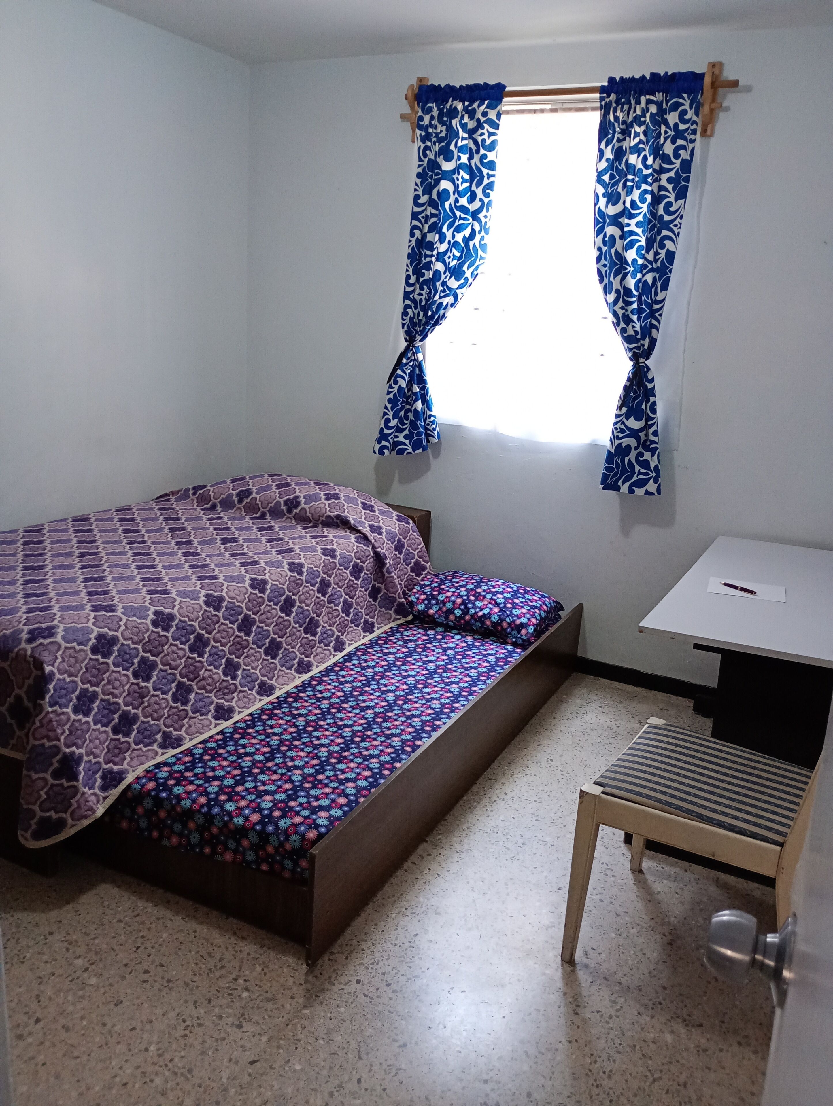 A small room with a bed, a desk, and a window with curtains.